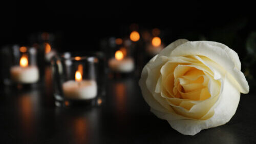 White rose and blurred burning candles on table in darkness, closeup with space for text. Funeral symbol